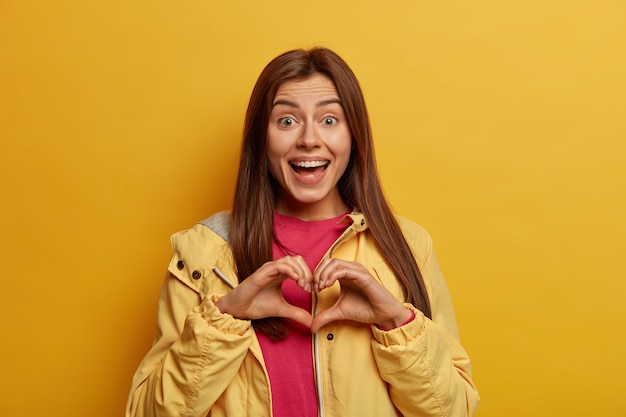 La giovane donna dai capelli scura ottimista modella il segno della mano del cuore sul petto, esprime amore, simpatia e affetto per la famiglia, guarda con gioia alla telecamera