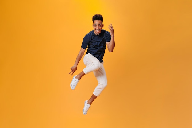 Оптимистичный темнокожий парень в стильном наряде прыгает на оранжевом фоне Брюнетка в черной футболке и белых штанах позирует на изолированном фоне