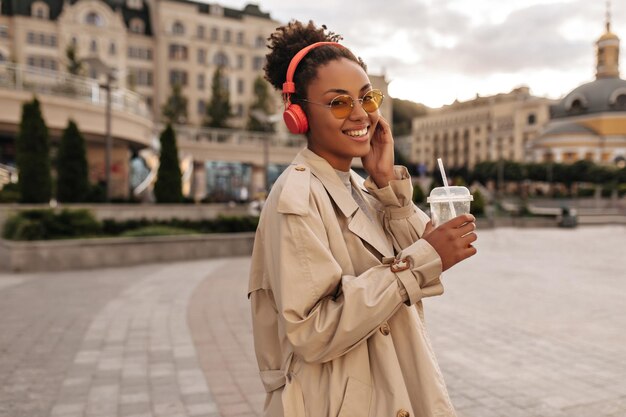 Optimistic darkskinned brunette woman in beige trench coat smiles listens to music in red headphones and holds orange juice glass outside
