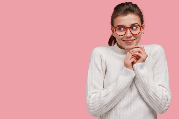 La giovane donna ottimista e sognante tiene le mani unite, concentrata da parte con un sorriso gentile, ha intenzione o piano per qualcosa di grandioso