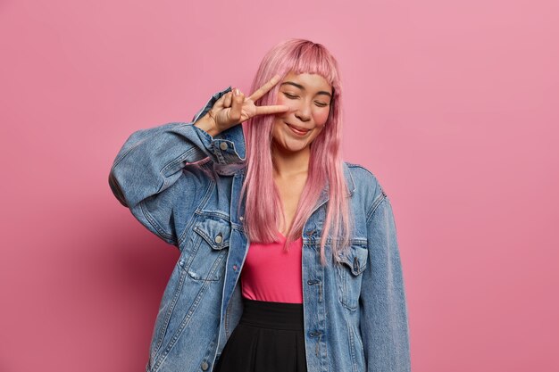 Optimistic energized long haired woman with eastern appearance, pink wig, makes peace gesture over eyes, smiles positively