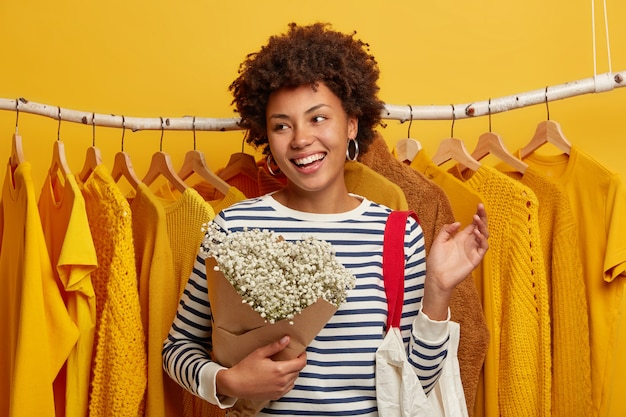 낙관적 인 여성 고객은 상점에서 쇼핑하고, 선반에있는 노란 옷에 꽃다발과 가방을 들고 포즈를 취하고, 활짝 웃으며 옆으로 집중합니다.