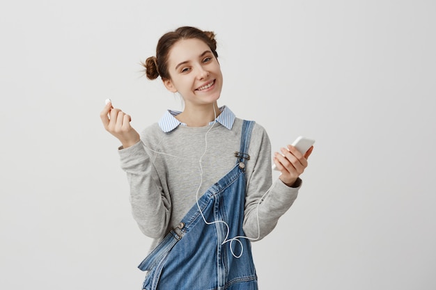 Optimistic girl in denim holding smartphone looking  with kind smile. Female DJ with nice appearance listening music via headphones taking pleasure. Technology concept