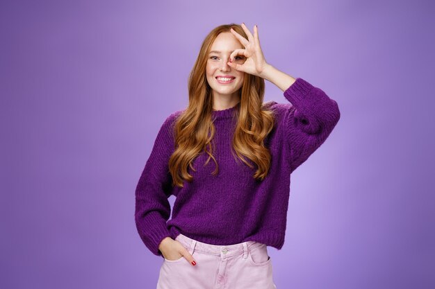 Оптимистичная девушка, смотрящая вдаль через нормальный жест и широко улыбаясь с обнадеживающей взволнованной улыбкой, стоя в восторге на фиолетовом фоне в свитере и рука в штанах.