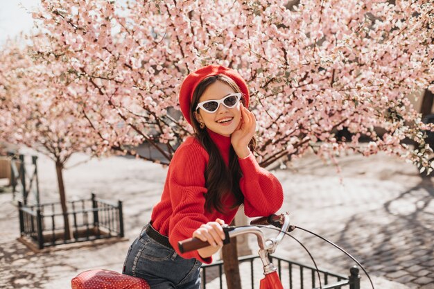 桜を背景に自転車に寄りかかって赤いセーターとメガネの楽観的な女の子。庭で笑っているスタイリッシュなベレー帽の魅力的な女性