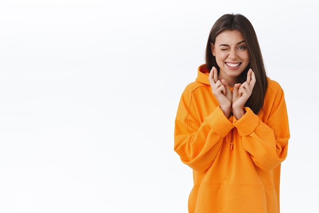 Optimistic good-looking modern girl in orange hoodie peeking at camera while cross fingers good luck and making wish, smiling hopeful