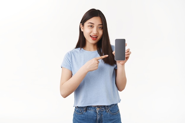 楽観的な格好良いアジアの女の子の黒髪は、興味深いアプリケーションゲームスタンド白い背景ショーソーシャルメディアプロファイルについて話すアプリホールドスマートフォンポインティング携帯電話画面を紹介します