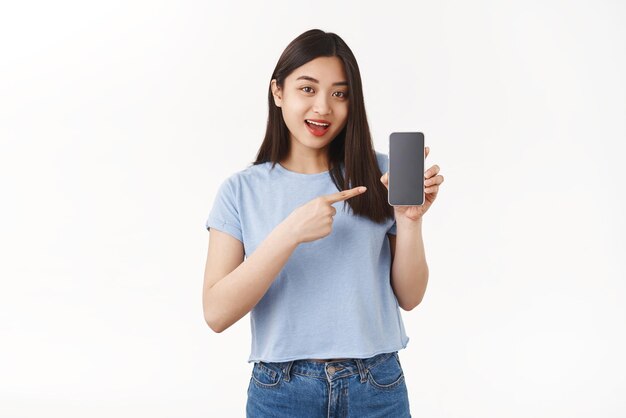 Оптимистичная красивая азиатская девушка с темными волосами представляет приложение, держит смартфон, указывая на экран мобильного телефона, говоря об интересном приложении, игровом стенде, белом фоне, показывает профиль в социальных сетях