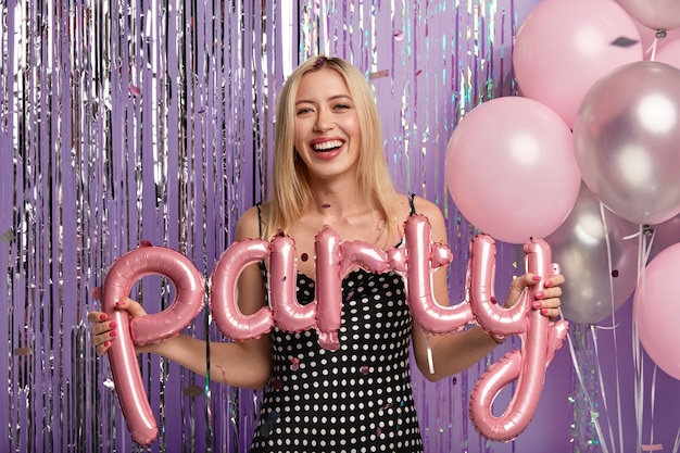 Optimistic happy blonde woman wears fashionable polka dot dress, makes photo with balloons on party