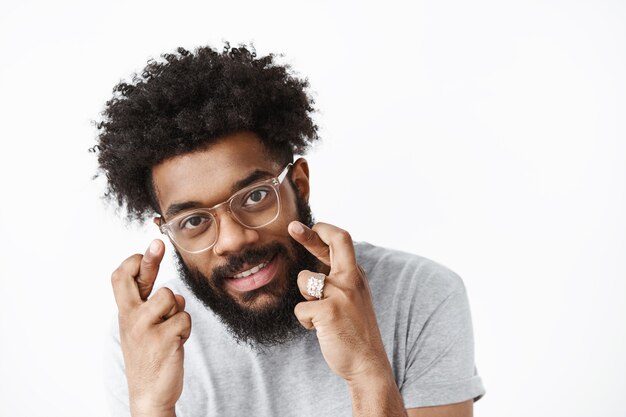 Optimistic and joyful african american bearded guy with afro hairstyle and glasses bending as crossing fingers for wishing luck smiling joyfully dreaming for pray come true over gray wall