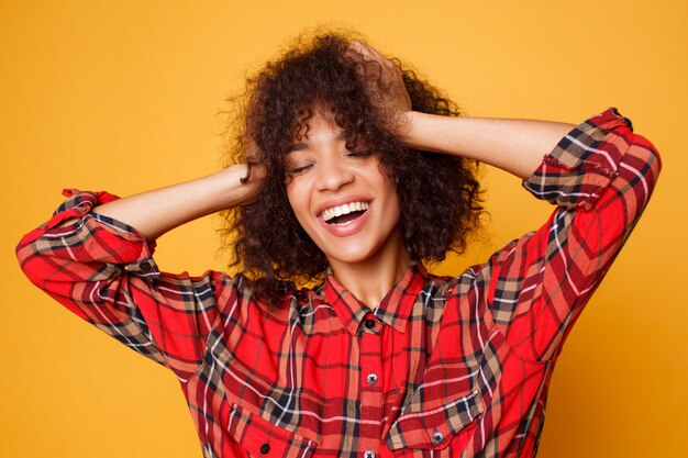 オレンジ色の背景のスタジオでポーズをとって楽観的な笑い黒人女性。人生を楽しむリラックスした巻き毛の女性モデル。