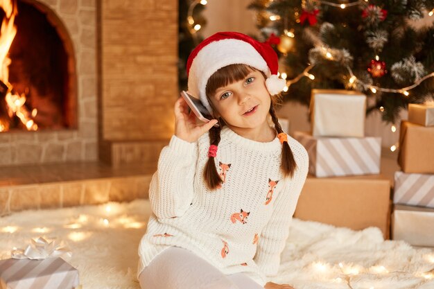 흰색 스웨터와 산타클로스 모자를 쓴 낙천적인 어린 소녀는 카메라를 보고, 축제 분위기를 갖고, 전화를 통해, 크리스마스 트리, 선물 상자, 벽난로 근처 바닥에 앉아 있습니다.