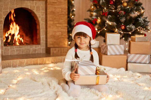 Optimistic little kid wearing white sweater and santa claus hat, sitting on soft carpet with stack of present boxes, posing in festive room with fireplace and Xmas tree.