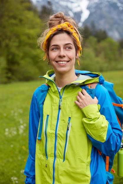 Viaggiatore adorabile ottimista con espressione felice, indossa una giacca a vento blu e verde, porta lo zaino