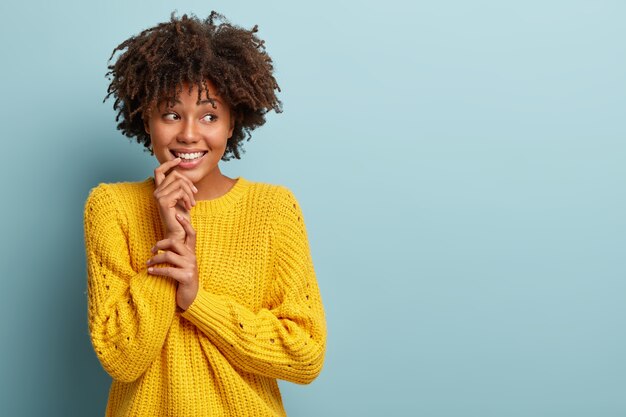 Optimistic lovely woman with curly hair, keeps hand near toothy smile, looks aside, has dreamy expression, feels shy, wears yellow casual jumper, stands against blue wall. Copy space right