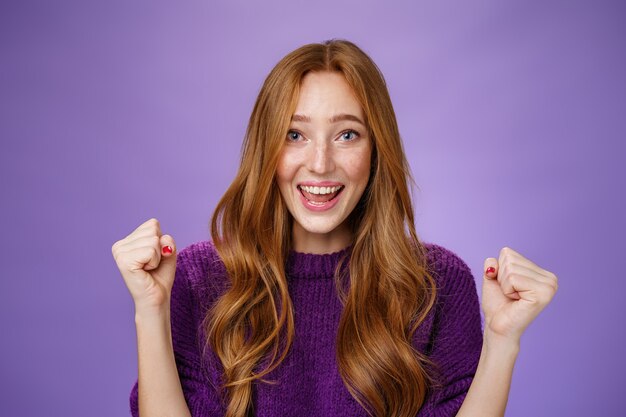Optimistic, lucky young attractive female student winning trip around european smiling broadly from success and delight clench fists in triumph and celebration gesture, happy to win over purple wall.