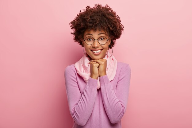 Optimistic smiling woman wears long sleeve jumper, keeps hands pressed together under chin