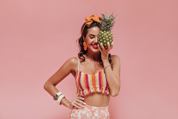 Optimistic trendy lady with orange bandana and red lips in fashionable summer clothes posing with pineapple and closed eyes..