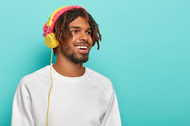 Optimistic unshaven male student has dreads, wears headphones on ears, has fun, listens favourite audio track, wears white clothes, looks aside, smiles pleasantly