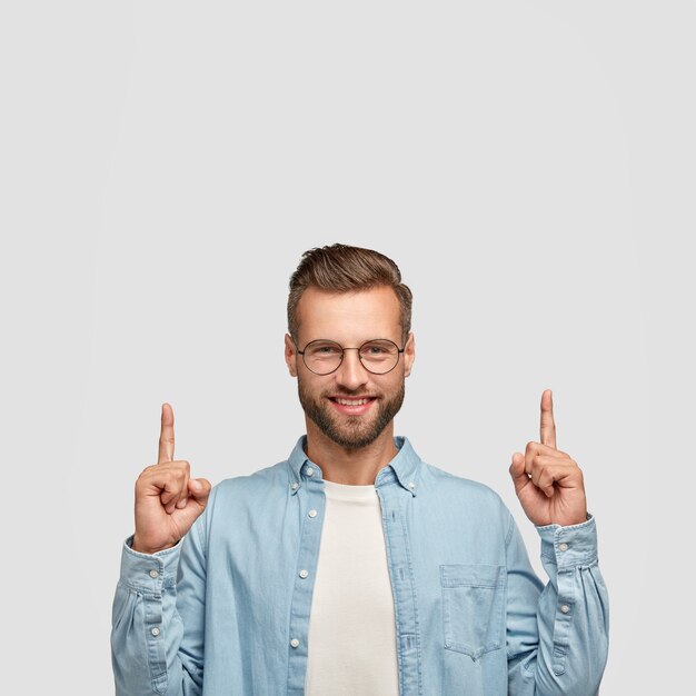 Optimistic unshaven young man has trendy haircut, cheerful expression, points upwards with both index fingers, being in good mood