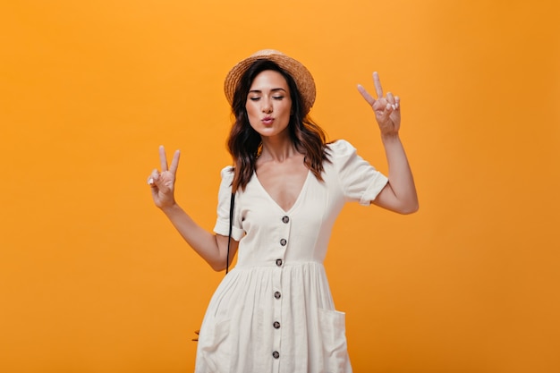 Optimistic woman in dress and hat shows signs of peace on orange background