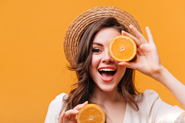 Optimistic woman in straw hat covers her eyes with orange and smiles while looking at camera.