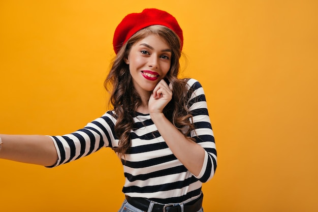 Optimistic woman in striped outfit smiles and makes selfie Beautiful lady in good mood with curly hair in red beret looking into camera