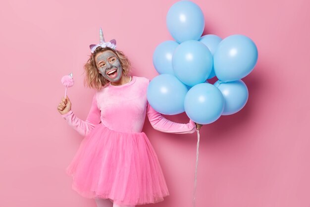 Optimistic woman with hairstyle applies clay facial mask for rejuvenation wears dress and unicorn headband dances carefree with bunch of blue balloons prepares for party isolated over pink background