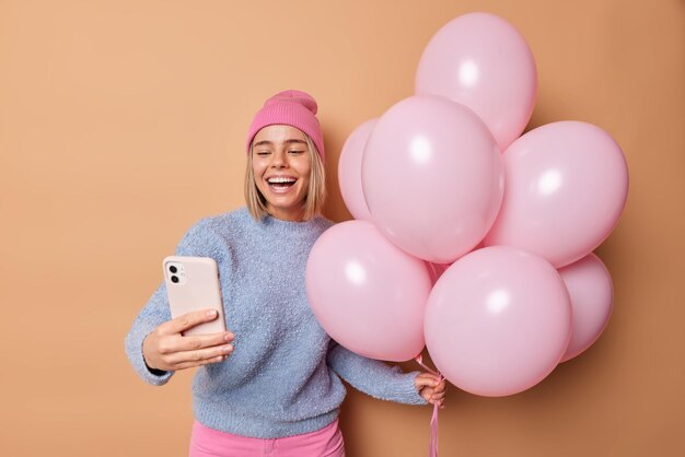 Optimistic young European woman laughs gladfully has upbeat mood takes selfie via modern smartphone wears hat and blue jumper celebrates anniversary holds bunch of inflated balloons poses indoor