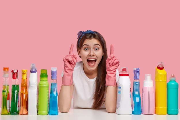 Optimistic young housemaid points upwards with both index fingers, wears headband and rubber gloves, shows something awesome on ceiling