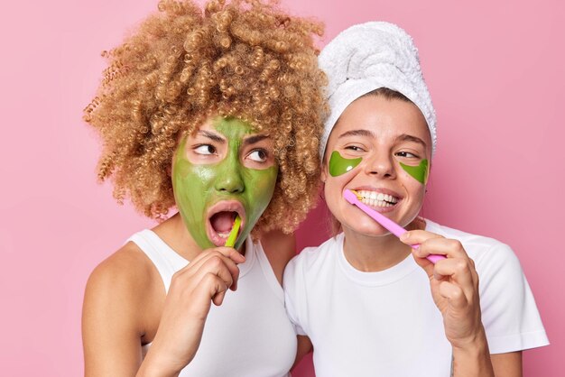 Oral hygiene concept Two women brush teeth with toothbrushes undergo daily hygiene routines apply beauty patches and nourishing mask wear towel on head casual t shirts isolated over pink backgroud