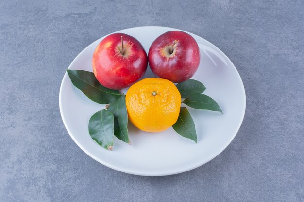 Orange and apples with leaves on plate on marble table.