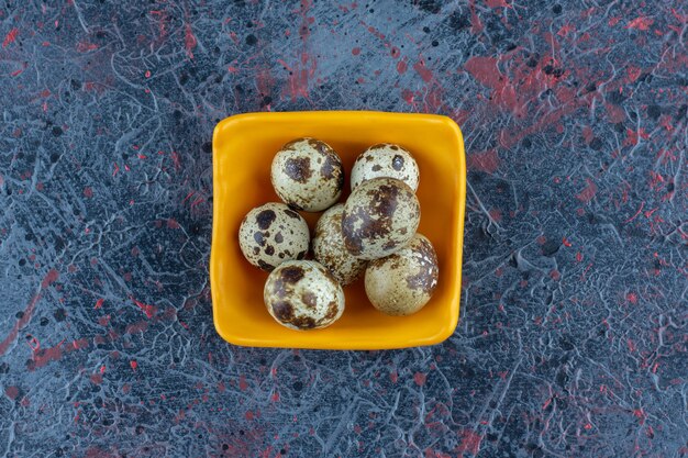 An orange bowl of quail eggs on marble table.