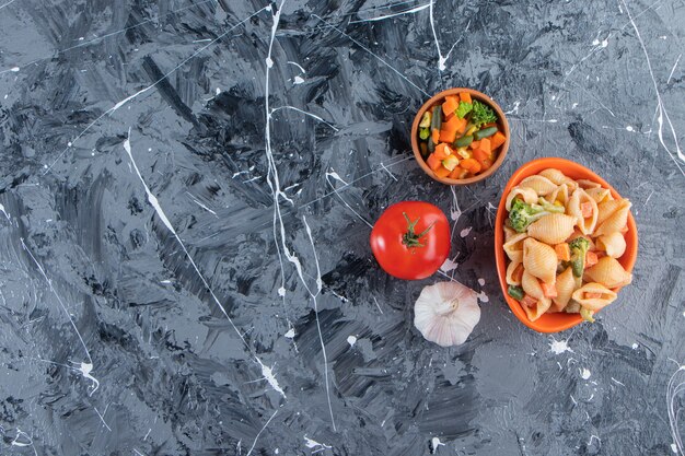 Orange bowl of tasty seashell pasta with vegetable salad on marble surface. 