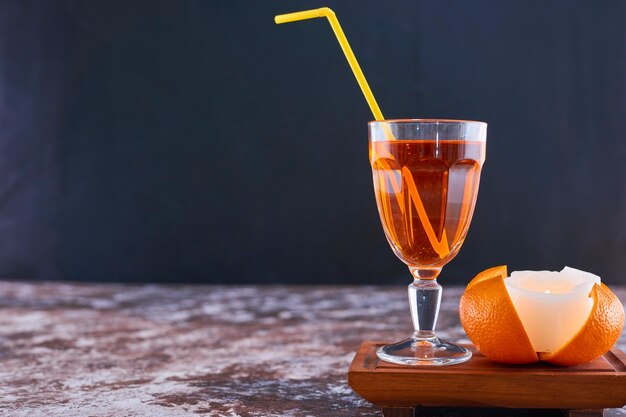 Orange and a glass of juice with yellow pipe on wooden platter on the marble
