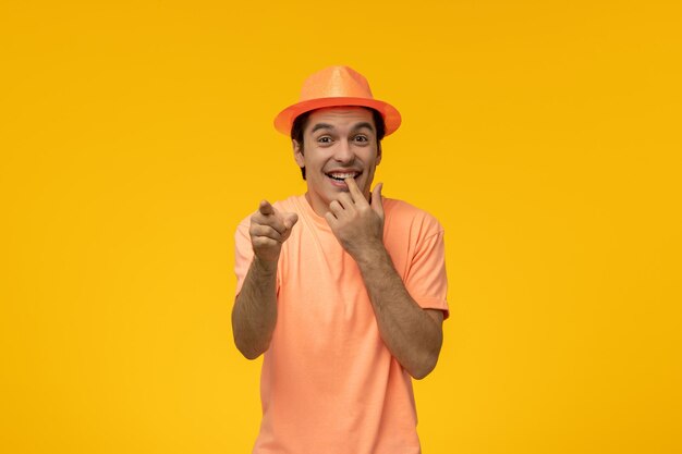 Оранжевая шляпа милый молодой парень в оранжевой рубашке со шляпой, кусающей палец и указывающей вперед