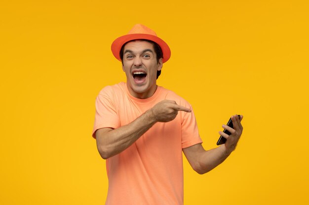 오렌지색 모자를 쓴 오렌지색 티셔츠를 입은 귀여운 젊은 남자가 모자를 쓰고 크게 웃고 전화를 보고 있다