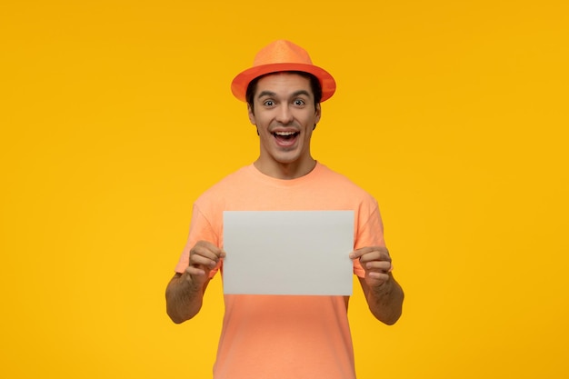 오렌지색 모자를 쓴 오렌지색 티셔츠를 입은 귀여운 젊은 남자가 웃고 있고 종이를 들고 있다