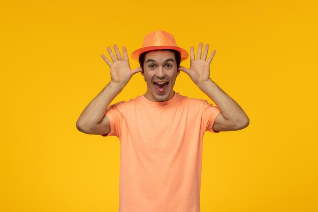 オレンジ色の帽子のハンサムなかわいい男は、帽子をいたずらして耳のジェスチャーをするオレンジ色のTシャツを着ています