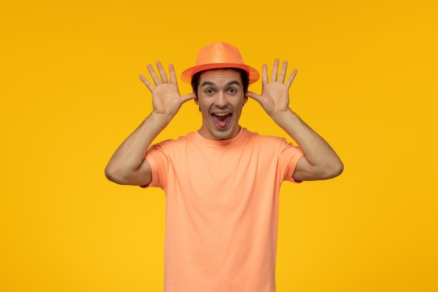オレンジ色の帽子のハンサムなかわいい男は、帽子をいたずらして耳のジェスチャーをするオレンジ色のTシャツを着ています