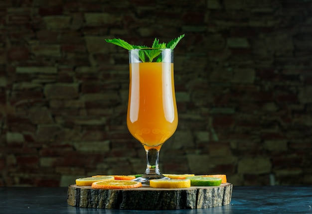 Orange juice in a goblet with mint, lime, lemon, orange, wooden board side view on blue and brick stone background