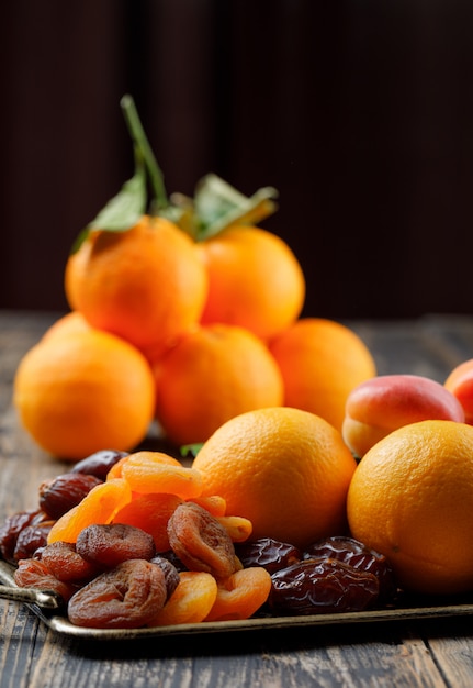 Oranges on branch with dates, dried and ripe apricots in tray side view on wooden table