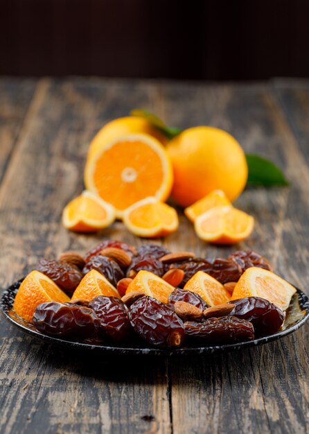 Oranges and slices in plate with dates, almonds and leaves on black and wooden table. vertical