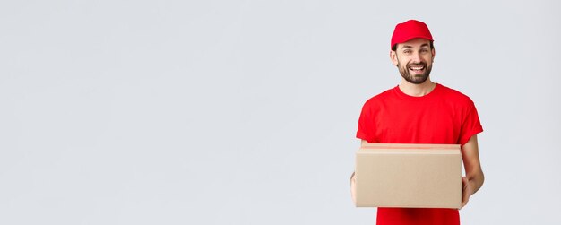 Order delivery, online shopping and package shipping concept. Friendly smiling courier in red uniform cap and t-shirt, handing out packages for customers. Employee bring parcel box, grey background.