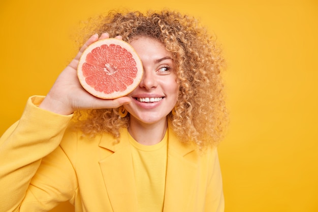 Organic diet concept. Pleased curly haired woman covers eye with grapefruit half smiles toothily going to make fresh juice or smoothie looks aside poses against yellow wall copy space area for text