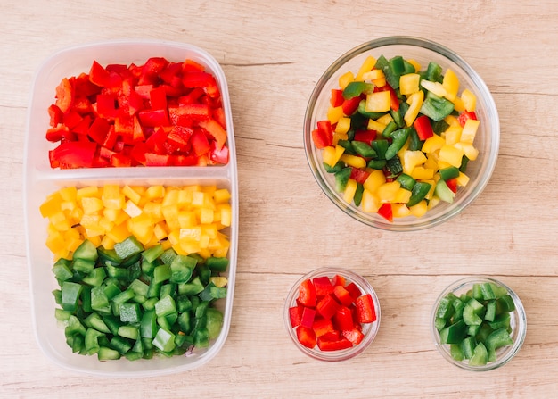 An organic fresh chopped red; green and yellow bell peppers in the tiffin box and glass bowls on wooden desk