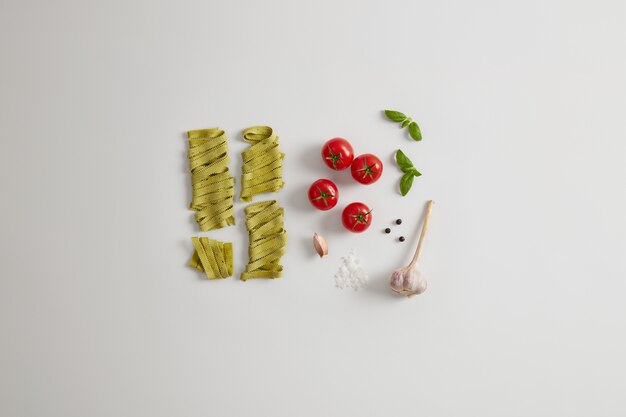 Organic green noodles with spinach, sea salt, fresh red tomatoes, garlic and basil leaves on white background. Preparing nourishing dish full of carbohydrates. Gluten free gourmet fettuccine