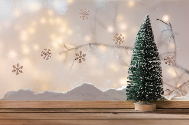 Ornament fir tree on wood table near bank of snow, plant twig, snowflakes and fairy lights