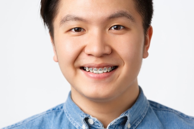 무료 사진 치열 교정 및 구강학 개념입니다. 웃고 있는 행복한 아시아 남성의 헤드샷, 치과 교정기를 보여주고, 진료소를 추천하고, 좋은 결과에 만족하고, 흰색 배경을 기쁘게 생각합니다.