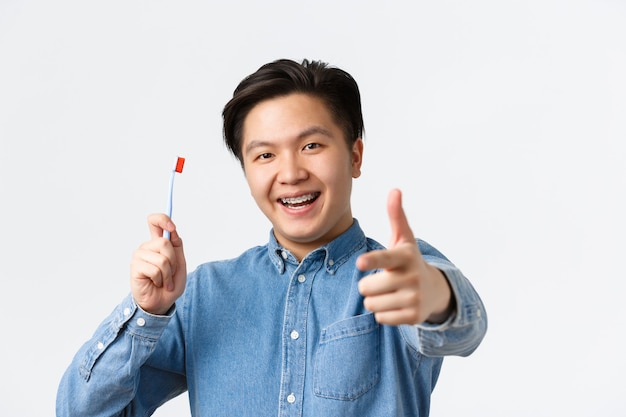 치열 교정, 치과 치료 및 위생 개념입니다. 행복하고 기뻐하는 아시아 남성의 클로즈업은 칫솔과 엄지손가락을 보여주는 구강 클리닉을 추천하며 치아 교정기를 관리하는 방법을 설명합니다.